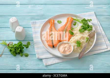 Frisch gebackene Lachssteaks mit Reis, Koriander und cremiger Sauce auf einem weißen Teller auf Holztisch mit Gabel und Serviette, horizobtaler Blick von oben, nicht Stockfoto