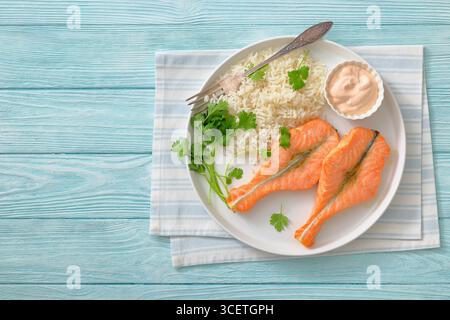 Gebackene Lachssteaks mit Reis, Koriander und cremiger Sauce auf einem weißen Teller auf Holztisch mit Gabel und Serviette, horizontaler Blick von oben, freier Platz, Stockfoto