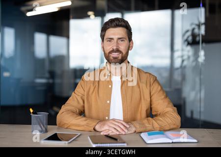 Porträt eines lächelnden Mannes, der an einem Schreibtisch in einem gut beleuchteten Büro sitzt, mit Notebooks und elektronischen Geräten auf dem Tisch, wodurch eine professionelle und zugängliche Atmosphäre entsteht. Stockfoto