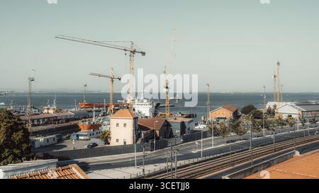 Geschäftiger Seehafen mit großen Kränen, Frachtschiffen, Lagerhäusern und Industriegebäuden entlang der Uferpromenade mit Blick auf das ruhige Meer mit weit entfernten Booten und Stockfoto