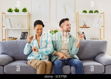 Glückliches junges, vielfältiges Paar in legeren Klamotten, das sich zu Hause auf dem Sofa im Wohnzimmer amüsiert. Stockfoto