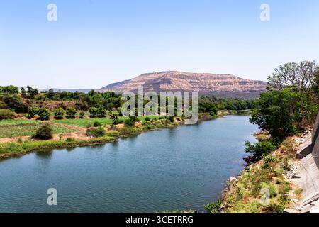 Ein ruhiger Fluss fließt durch üppiges Grün mit einem Berg im Hintergrund unter einem klaren blauen Himmel. Stockfoto