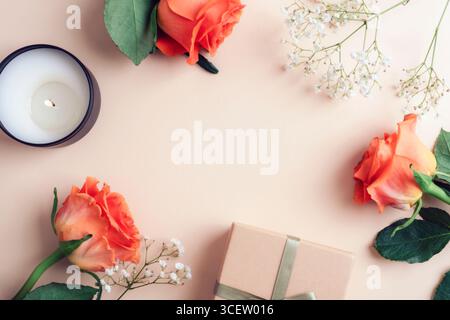 Rahmen aus drei scharlachroten Rosen, Geschenkbox und Kerze auf beigefarbenem Hintergrund. Weihnachtsgrußkarte, Kopierraum. Stockfoto