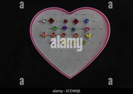 Niedliche Kinderringe mit Zeichentrickmotiven in einer herzförmigen Box auf schwarzem Hintergrund angeordnet Stockfoto