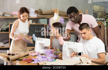 Männlicher Lehrer mittleren Alters hilft Schülern beim Nähen, während andere mit Textilien arbeiten Stockfoto