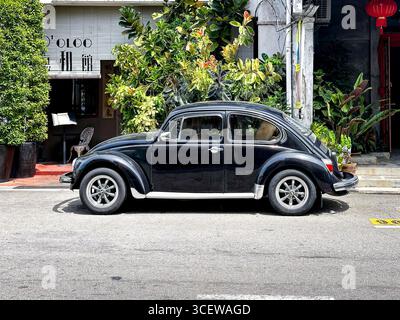 Seitenansicht des schwarzen Vintage Volkswagen Käfers Typ 1 auf der Straße mit üppigen grünen Pflanzen und Gebäudeeingang im Hintergrund Malacca malaysia Stockfoto
