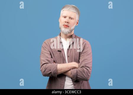 Junger Albino-Mann posiert mit gekreuzten Armen und schaut mit Ernsthafter Gesichtsausdruck bei der Kamera auf türkisfarbenem Hintergrund Stockfoto