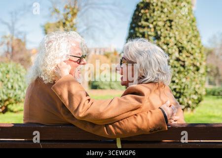 Glückliches Seniorenpaar, das sich liebevoll umarmen und lachen lässt, während es auf einer Bank sitzt und einen besonderen romantischen Moment genießt, der ihre hervorhebt Stockfoto