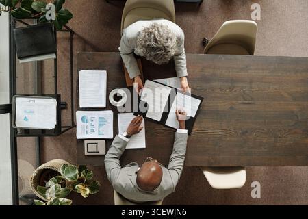 Kaukasische Seniorin und schwarzer Mann mittleren Alters sitzen am Holztisch und prüfen gemeinsam Geschäftsdokumente, analysieren Diagramme und Diagramme, arbeiten im Büro zusammen, Ansicht von oben Stockfoto
