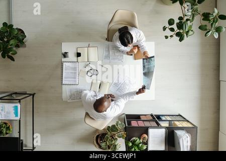Schwarzer Mann mittleren Alters und junge Erwachsene asiatische Frau sitzen am Schreibtisch und überprüfen medizinische Dokumente und untersuchen Röntgenbilder zusammen im Büro, aus Sicht von oben Stockfoto
