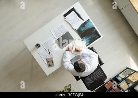 Kaukasier mittleren Alters sitzt am Schreibtisch, liest medizinische Dokumente und schreibt in Notizbuch, analysiert Röntgenbilder, zeigt einen organisierten Arbeitsbereich mit Papieren und Ordnern Stockfoto