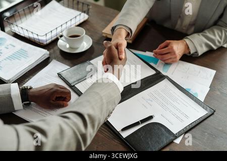 Kaukasische Frau mittleren Alters und schwarzer Mann mittleren Alters schütteln die Hände über Vertragsdokumente auf dem Schreibtisch, beide tragen Geschäftskleidung, Draufsicht zeigt professionelle Zustimmung Stockfoto