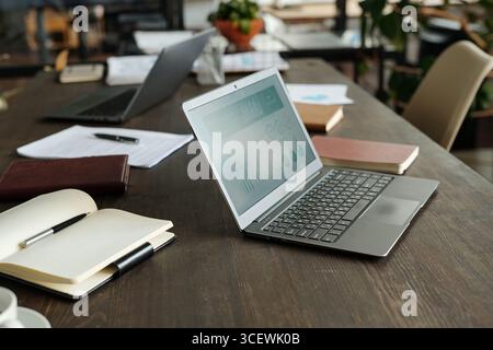 Draufsicht mit offenem Laptop, auf dem Geschäftsanalysen auf einem Holztisch dargestellt werden, umgeben von Notizbüchern, Dokumenten, Stift und einem anderen Laptop, Arbeitsbereich für Büroarbeiten oder Remote-Meetings Stockfoto