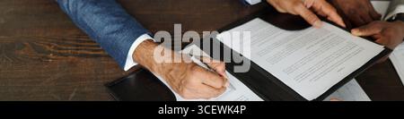 Kaukasischer Mann mittleren Alters unterschreibt Vertrag, während schwarzer Mann mittleren Alters auf ein Dokument auf einem Holztisch zeigt, von oben mit Händen und Papieren während des Geschäftsprozesses Stockfoto
