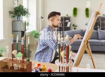 Beschäftigter Mann, der kreative Kunstbilder auf Staffelei malt, Künstler, der Pinsel hält, um im Studio zu Hause zu malen Stockfoto