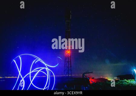 Kreative Langzeitbelichtung von Lichtspuren vor einem rot beleuchteten Telekommunikationsturm auf einem Hügel, unter einem klaren Sternenhimmel und glühenden Lichtern der Stadt. Stockfoto