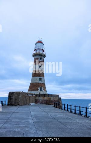 Blick auf einen hohen Leuchtturm aus Stein mit roten und weißen Streifen an der Küste mit Blick auf das Meer unter einem bewölkten Himmel Stockfoto