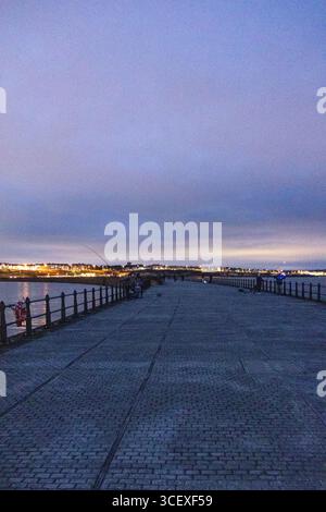 Perspektivische Aussicht auf einen langen Steg mit Geländern, der sich zum Meer und zum Leuchtturm unter einem dramatischen Abendhimmel erstreckt Stockfoto