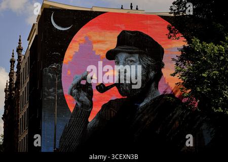 Buntes Wandgemälde eines bärtigen Mannes mit einer Pfeife vor einem roten Himmel und einer Halbmond, das auf einem hohen Gebäude neben Bäumen und Wolken in Amsterdam zu sehen ist. Stockfoto