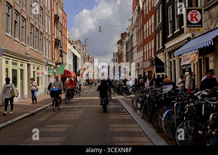Die Menschen radeln entlang einer Straße, die von Geschäften und Fahrradreihen, Ziegelfassaden und hängenden Lichtern gesäumt ist, unter Wolken auf der Haarlemmerdijk in Amsterdam. Stockfoto