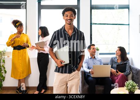 Porträt eines jungen Managers, der einen Laptop hält und mit Kollegen lächelt, die Laptop und Tablet benutzen, um im Hintergrund bei einem Startup-Unternehmen zu diskutieren Stockfoto
