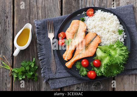Gebackene Lachssteaks mit Reis, Salat, Tomaten auf einem schwarzen Teller auf rustikalem Holztisch mit Gabel und Serviette, horizontaler Blick von oben, flacher Lay, Clo Stockfoto
