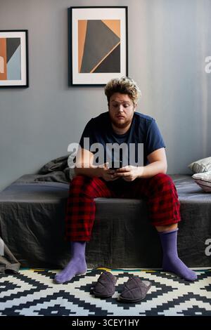 Vertikale Aufnahme eines jungen kaukasischen Mannes mit Schlafanzug, der morgens auf dem Bett sitzt und etwas auf dem Smartphone ansieht Stockfoto