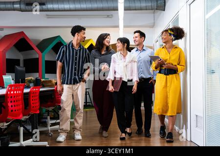 Multiethnische Geschäftsteams gehen und unterhalten sich gemeinsam in einem modernen Bürokorridor, arbeiten zusammen und diskutieren Arbeitsangelegenheiten in einem dynamischen Startup en Stockfoto