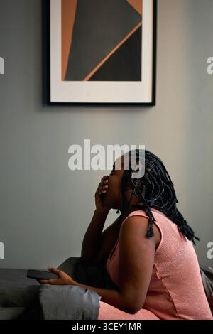 Vertikale Seitenansicht Porträt der verschlafenen jungen Schwarzen Frau, die morgens auf dem Bett sitzt und das Smartphone hält und gähnt Stockfoto
