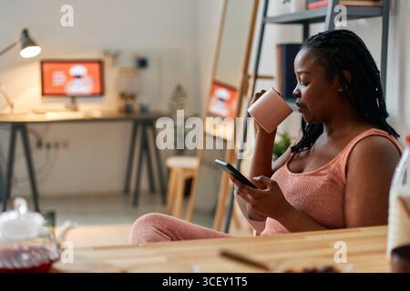 Seitenansicht Porträt einer jungen Schwarzen Frau, die den Morgen zu Hause entspannt sitzt und Tee trinkt und sich etwas im Internet auf dem Smartphone ansieht Stockfoto