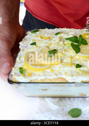 Italienisches Tiramisu. Hand hält ein Tablett mit Zitronen-Tiramisu mit Marienfinger und Mascarponcreme auf einem weißen Tisch, frische Minzdekoration. Sommerdessert. Stockfoto