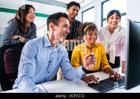 Verschiedene Kollegen treffen sich um einen Computer herum, arbeiten zusammen und lächeln, während sie in einem hellen, modernen Büro zusammenarbeiten und Ideen und Fachwissen austauschen Stockfoto