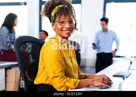 Junge Geschäftsfrau mit gelbem Hemd und Brille, lächelnd, während sie in einem modernen Büro mit Kollegen arbeitet, die in t zusammenarbeiten Stockfoto