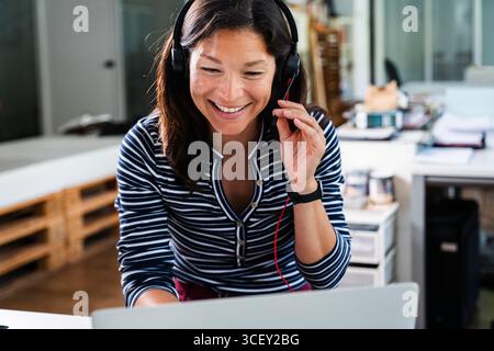 Glückliche Callcenter-Mitarbeiterin, die ein Headset trägt und am Laptop arbeitet, unterstützt und Kundenanfragen in modernen Startup-Büros beantwortet Stockfoto