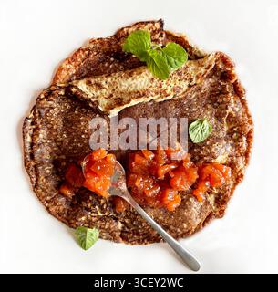 Dünne Vollkorncrepé. Ein Teller mit hausgemachten Pfannkuchen-Crepes mit Marmelade isoliert auf einem weißen Tisch. Direkt darüber. Stockfoto