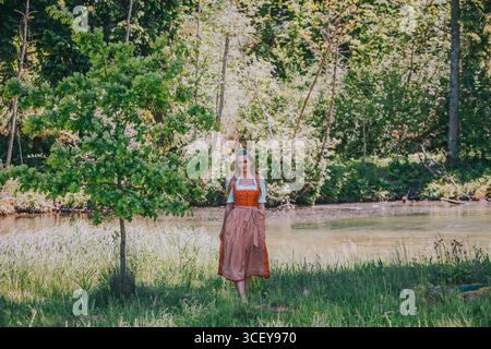 Eine junge Frau mit langen Haaren, gekleidet in einem traditionellen roten Dirndl mit weißer Bluse und roter Schürze, steht inmitten einer idyllischen Naturlandschaft Stockfoto