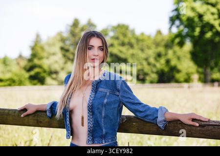 Eine junge Frau mit langen blonden Haaren posiert draußen. Sie trägt eine offene Jeansjacke und Jeansshorts, die zur Seite schauen. Im Hintergrund Stockfoto