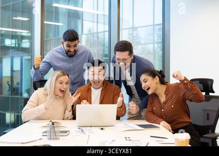 Eine Gruppe von fünf Fachleuten drückt Freude und Begeisterung aus, während sie auf einen Laptop-Bildschirm schauen und Teamarbeit, Erfolg und Feier in einem modernen Büro mit großen Fenstern vermitteln. Stockfoto
