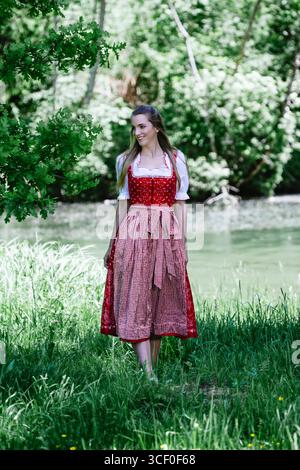 Eine junge Frau in Einem traditionellen Rot-weiß-karierten Dirndl läuft auf Einem schmalen Pfad am Ufer Eines Flusses. Sie Lächelt Und Schaut Zur Seite. T Stockfoto