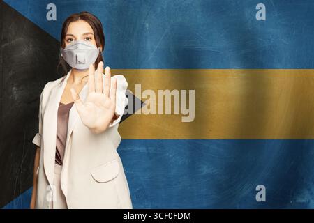 Selbstbewusste Frau in medizinischer Maske zeigt Stop Geste geöffnete Handfläche gegen Flagge von Bahamas Hintergrund Stockfoto