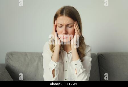Erschöpfte, müde junge Frau, die den Kopf berührt, unter Kopfschmerzen, Stress, Müdigkeit, Schläfenschmerzen, Migräne, lautes Geräusch, Spannung auf der Couch Stockfoto