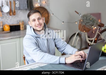 Ein freiberuflicher Kerl sitzt am Küchenschreibtisch mit Laptop. Bärtiger Freelancer, männlich, der von zu Hause aus arbeitet. Mann chatten mit Freunden online von zu Hause aus über den pc. Stockfoto
