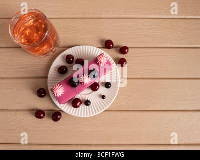 Rosafarbenes Gebäck mit Heidelbeeren und rosa cremefarbenen Blüten auf einem weißen Teller, umgeben von Kirschen und einem Orangengetränk auf einer rustikalen Holzoberfläche Stockfoto