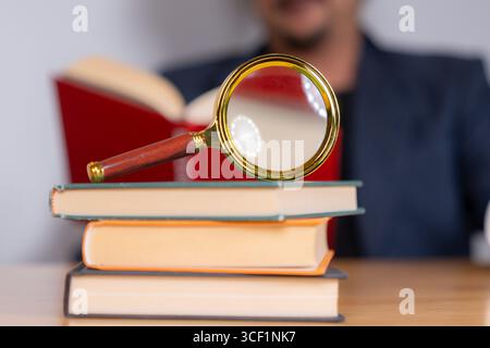 Ein Erwachsener sitzt und liest ein Buch sorgfältig mit einer Lupe, wobei er sich auf Details zum Studium, Wissen oder Forschung konzentriert Stockfoto