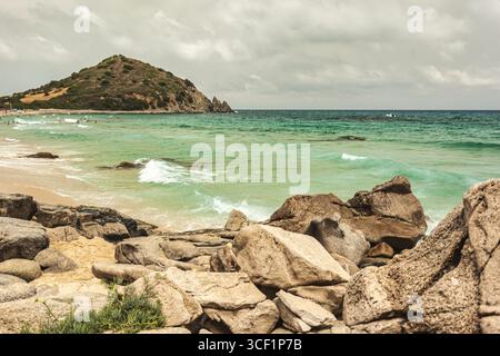 Sanfte Wellen, die über Felsen am Strand von monte turnu, italien, mit einem bewölkten Himmel und Touristen, die das Meer genießen Stockfoto