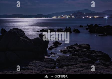 Ruhige Mondszene am Strand des Capo Ferrato in sardinien, italien, mit Felsen, ruhigem Meer und fernen Lichtern der Stadt bei Nacht Stockfoto