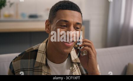 Positiver afroamerikanischer Mann männlicher Kerl genießt gute Kommunikation mit Smartphone im Wohnzimmer sprechendes Handy lächelndes Gespräch mit Stockfoto