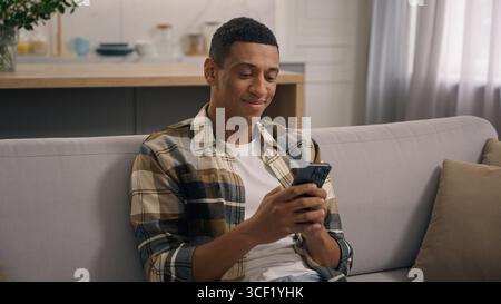 Glückliches Lachen positiv entspannter afroamerikanischer Mann zu Hause mit Handy Smartphone-Anwendung lächelnd drinnen im Wohnzimmer Lächeln Mann männlich Stockfoto