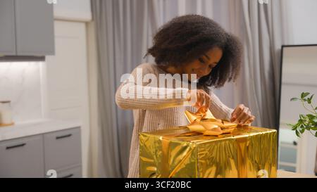 Sorglos aufgeregt Afroamerikanerin Käuferin Mädchen weibliche Klientin kommt nach Hause und hält Lieferservice Goldbox Geburtstagsgeschenk lächelnd Stockfoto