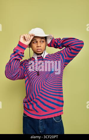 Ein stylischer junger Schwarzer posiert selbstbewusst in einem einzigartigen gestreiften Sweatshirt vor grünem Hintergrund. Stockfoto